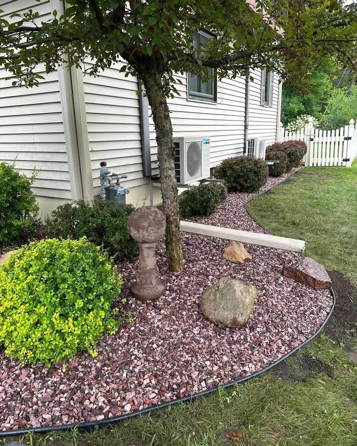 Curved landscape bed of red rock with shrubs and a birdbath under a tree, steel edging along the lawn