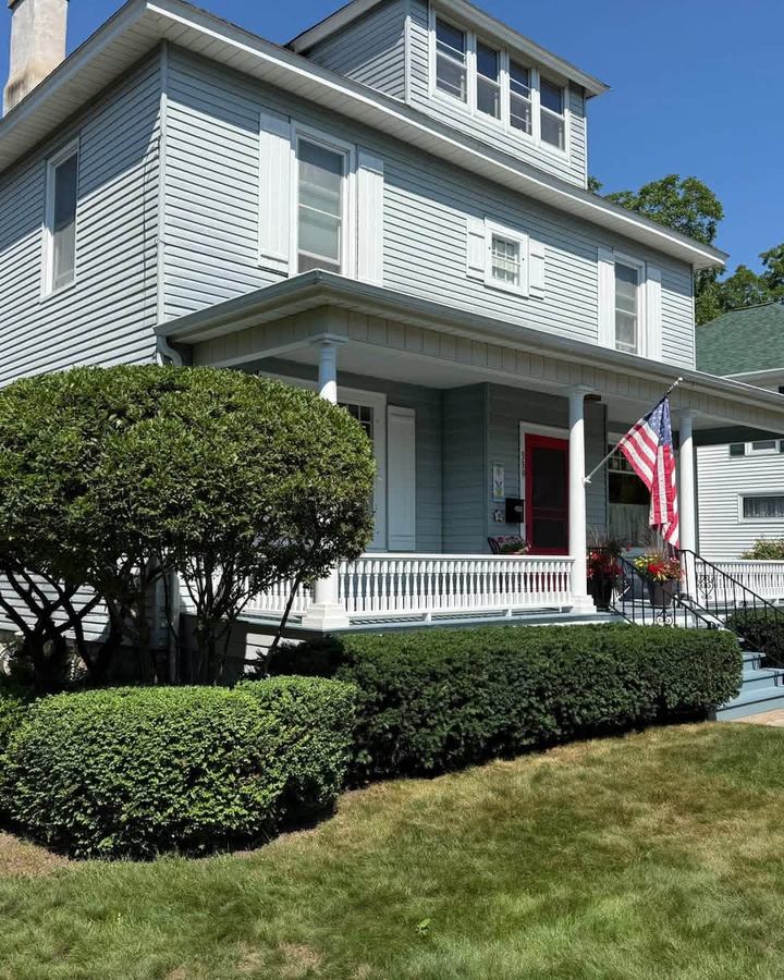 Two-storey grey house with a porch, an American flag and rounded hedges over a fresh cut lawn