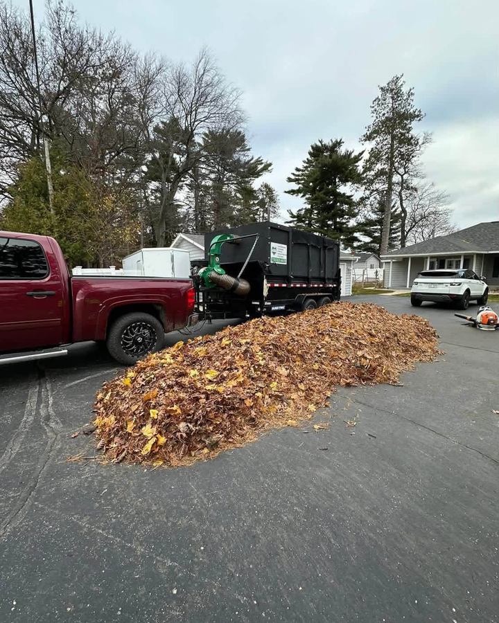Large pile of raked leaves on a driveway beside a red pickup and a dump trailer